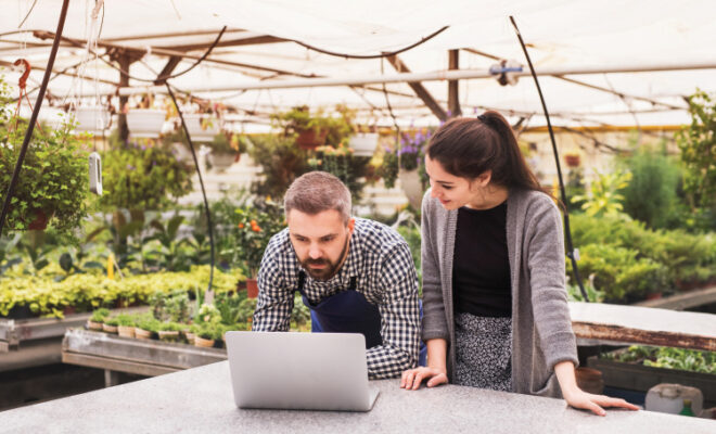 Young gardeners with laptop in a large greenhouse with pots of seedlings and plants clearning their inbox.